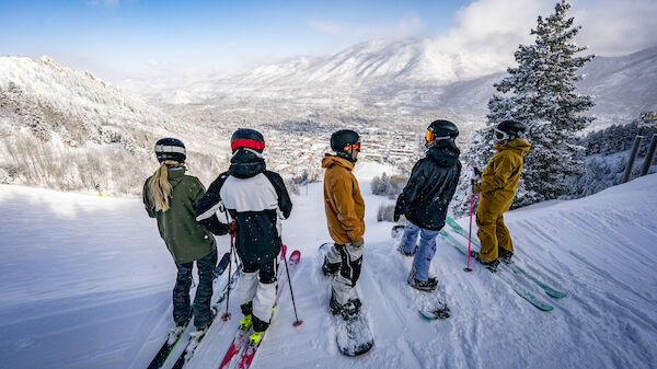 A group of people in winter gear stands on a snow-covered mountain, with skis and snowboards, looking at a scenic view of distant mountains.