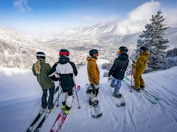 A group of people in winter gear stands on a snow-covered mountain, with skis and snowboards, looking at a scenic view of distant mountains.