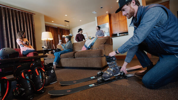 A person is preparing skiing gear while others sit on a couch in a cozy living room, suggesting a ski trip or winter vacation.