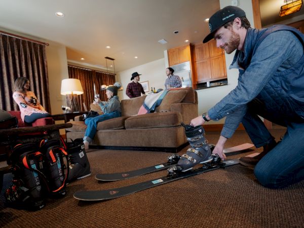 A person is preparing skiing gear while others sit on a couch in a cozy living room, suggesting a ski trip or winter vacation.