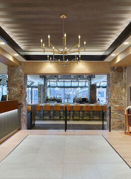 A modern hotel lobby with a reception desk on the left, a glass-fronted entrance area in the center, seating areas on the right, warm wood and stone accents, and a chandelier overhead, all under soft ambient lighting.