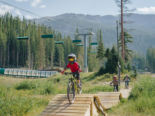 Three cyclists are riding on a mountain trail with elevated wooden sections surrounded by greenery and trees, and mountains in the background.