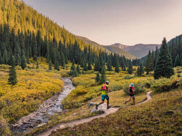 Two people are trail running with a dog in a scenic mountainous area with a stream, surrounded by evergreen trees and autumn foliage, during sunset.