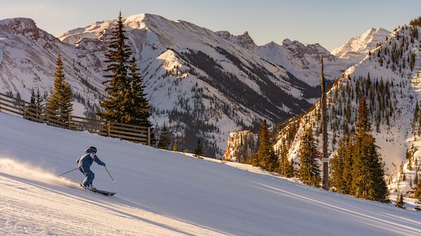 A skier glides down a snowy mountain slope with pine trees and rugged, sunlit alpine peaks in the background.