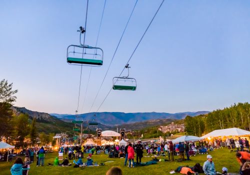 People gathered outdoors at an event with tents, surrounded by mountains. Ski lifts overhead, grass, and clear sky visible.