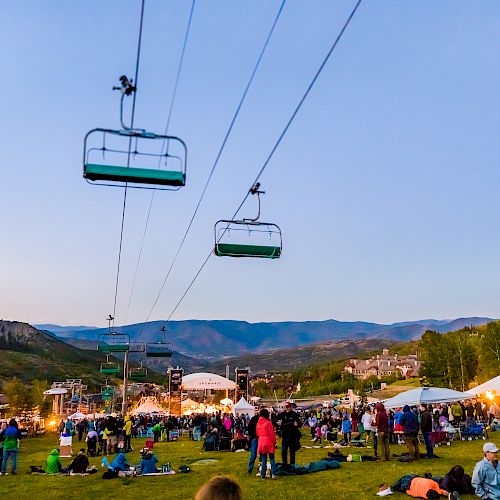 People gathered outdoors at an event with tents, surrounded by mountains. Ski lifts overhead, grass, and clear sky visible.