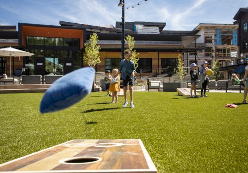 People playing cornhole in a grassy outdoor area with buildings in the background, on a sunny day.