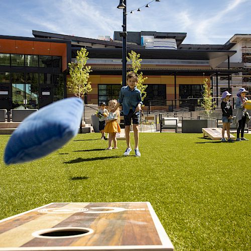 People playing cornhole in a grassy outdoor area with buildings in the background, on a sunny day.