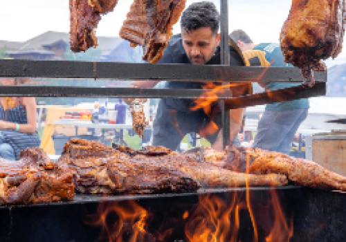 A man is cooking large cuts of meat over an open flame at an outdoor barbecue or grilling event, with people seated in the background.