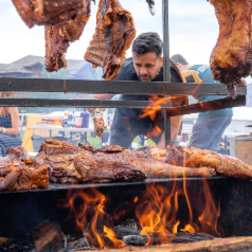 A man is cooking large cuts of meat over an open flame at an outdoor barbecue or grilling event, with people seated in the background.