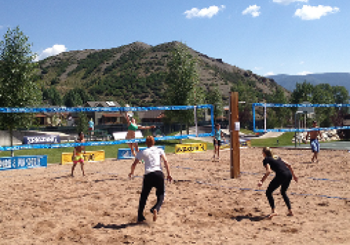 People playing beach volleyball on a sunny day with mountains and trees in the background. The scene looks lively and outdoorsy.