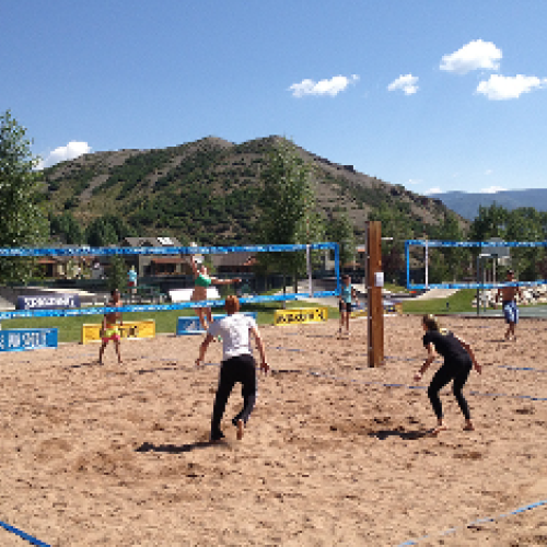 People playing beach volleyball on a sunny day with mountains and trees in the background. The scene looks lively and outdoorsy.