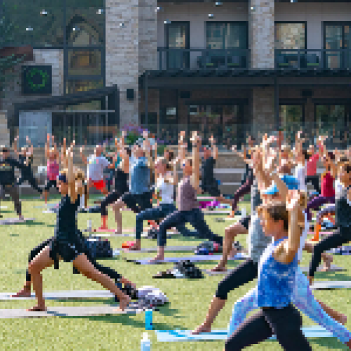 A group of people is participating in an outdoor yoga class on a grassy area in front of a building, practicing various poses together.