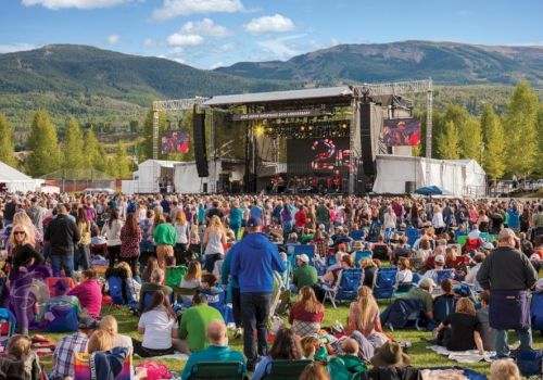 A large outdoor concert is taking place with a big stage, screens, and a crowd of people in front, surrounded by mountains and trees under a blue sky.