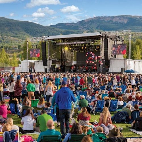 A large outdoor concert is taking place with a big stage, screens, and a crowd of people in front, surrounded by mountains and trees under a blue sky.