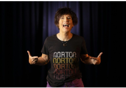 A person stands in front of a dark curtain, wearing a T-shirt that reads 