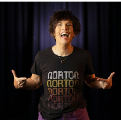 A person stands in front of a dark curtain, wearing a T-shirt that reads 