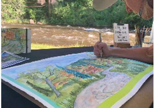 An artist, wearing a hat, is working on a colorful landscape painting at a riverside outdoor setting. Brushes and a phone are on the table.