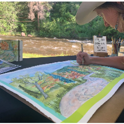 An artist, wearing a hat, is working on a colorful landscape painting at a riverside outdoor setting. Brushes and a phone are on the table.