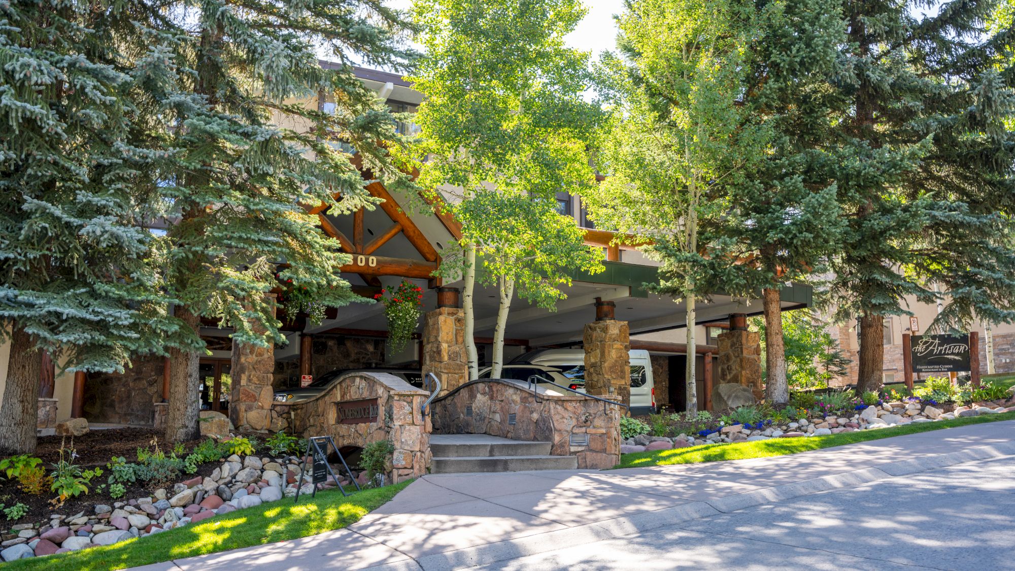 A scenic outdoor setting featuring a rustic wooden and stone structure, surrounded by lush trees and landscaped greenery, with a pathway leading up.