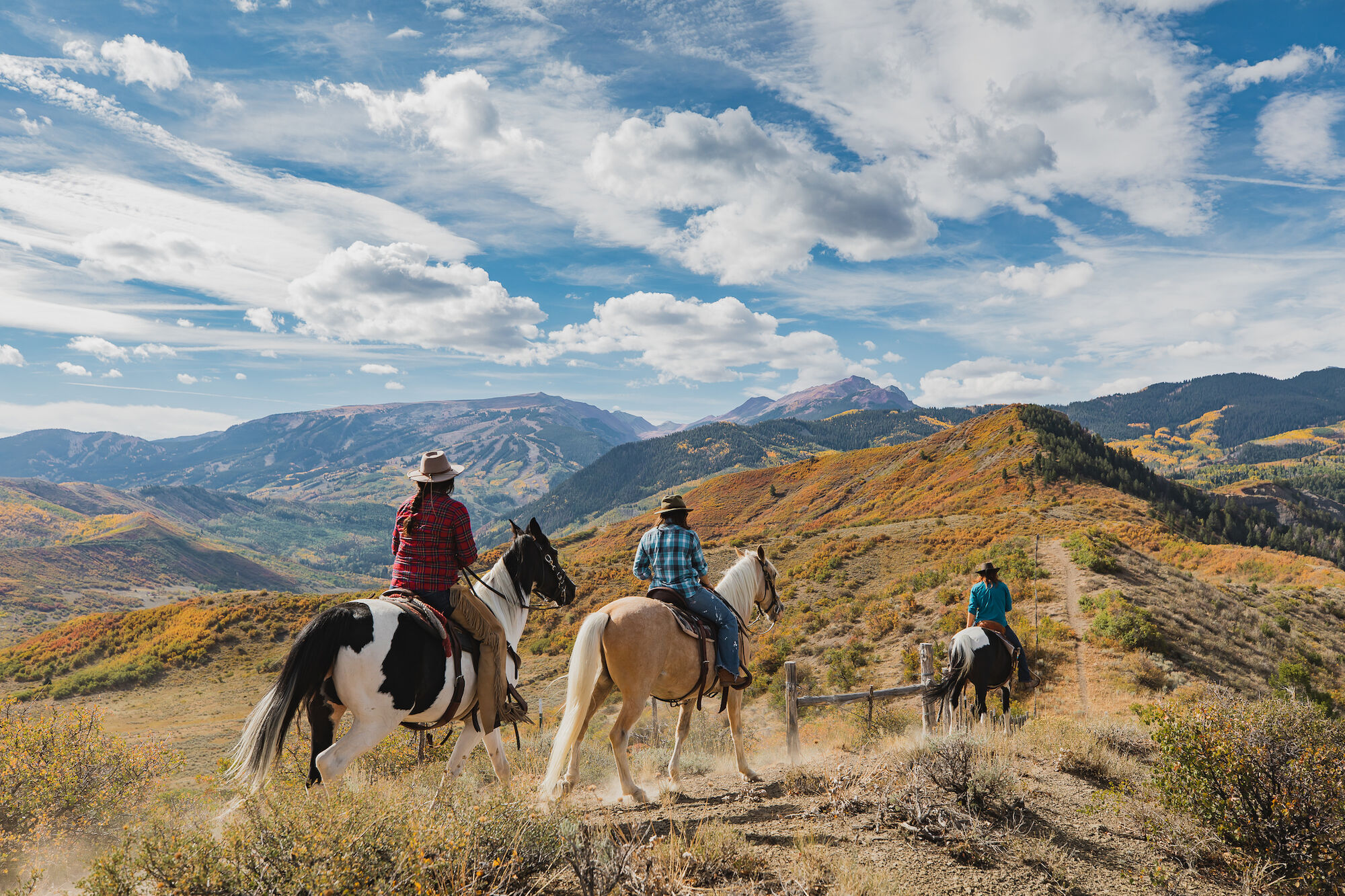 Three people are horseback riding through a scenic mountainous landscape under a partly cloudy sky.