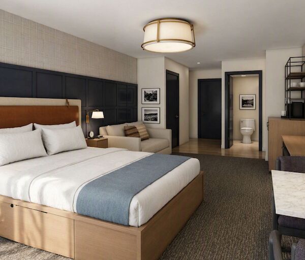 A modern hotel room with a large bed, blue upholstered headboard, nightstands, a desk, seating area, and a hallway leading to a bathroom.