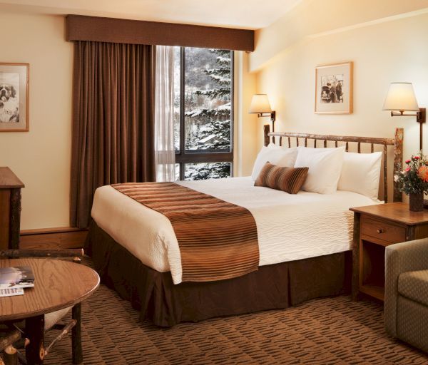A cozy hotel room with a bed, nightstands, lamps, a TV, a small table with magazines, artwork on the wall, and a window with tree views.