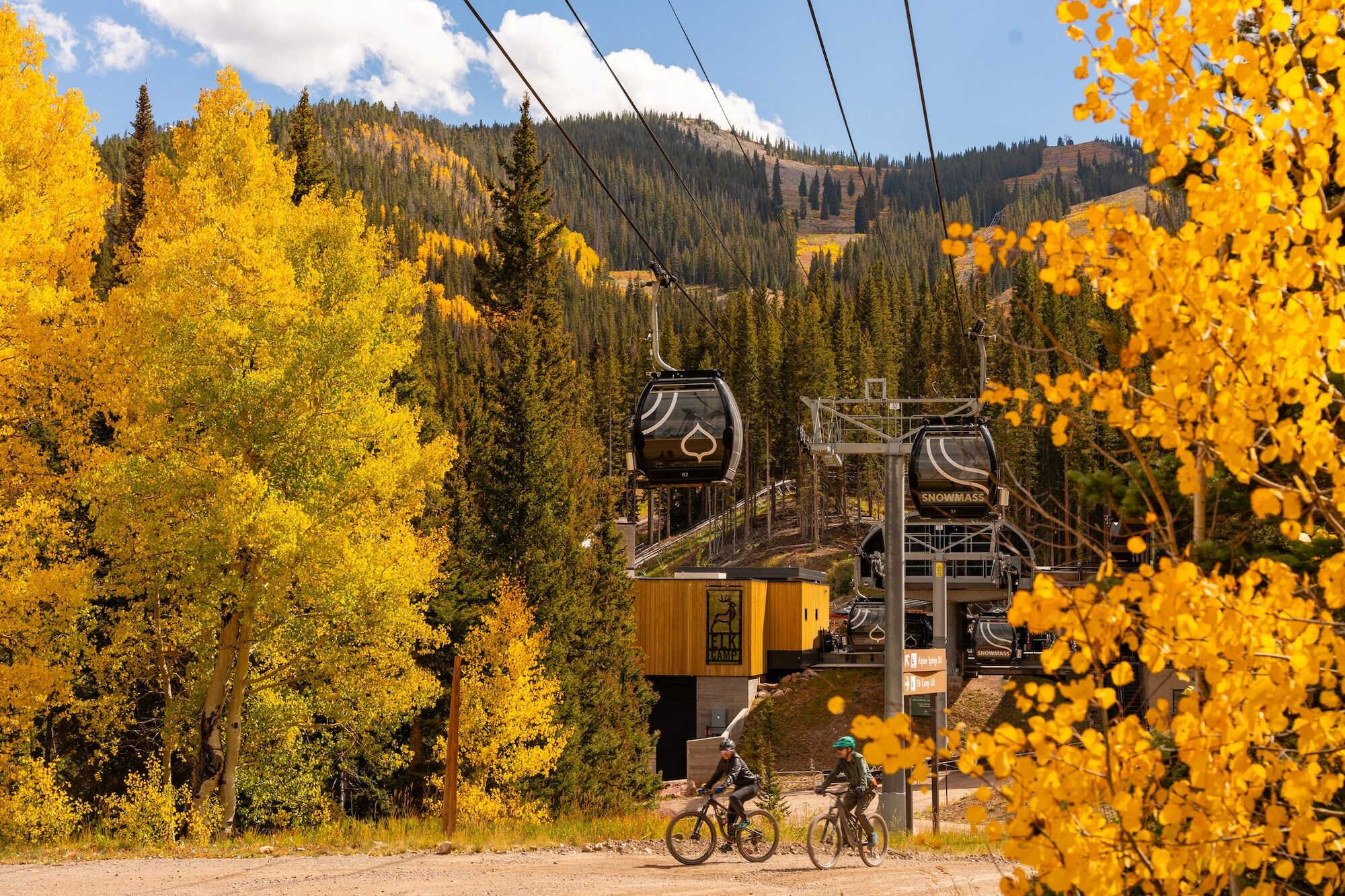 A scenic autumn landscape features a ski lift with gondolas, surrounded by vibrant yellow trees and two bicycles parked nearby.