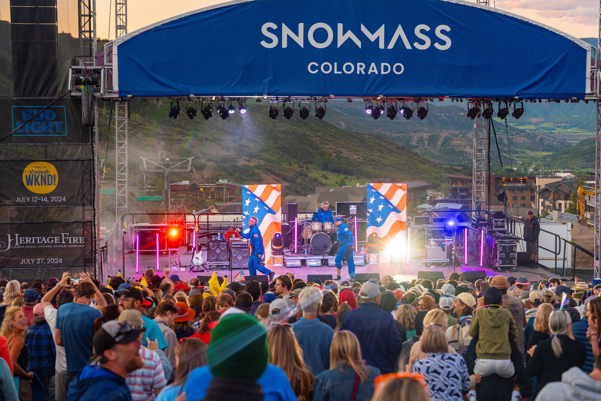 A concert at Snowmass, Colorado, with a large crowd, lively stage with lights, and performers with American flag visuals.