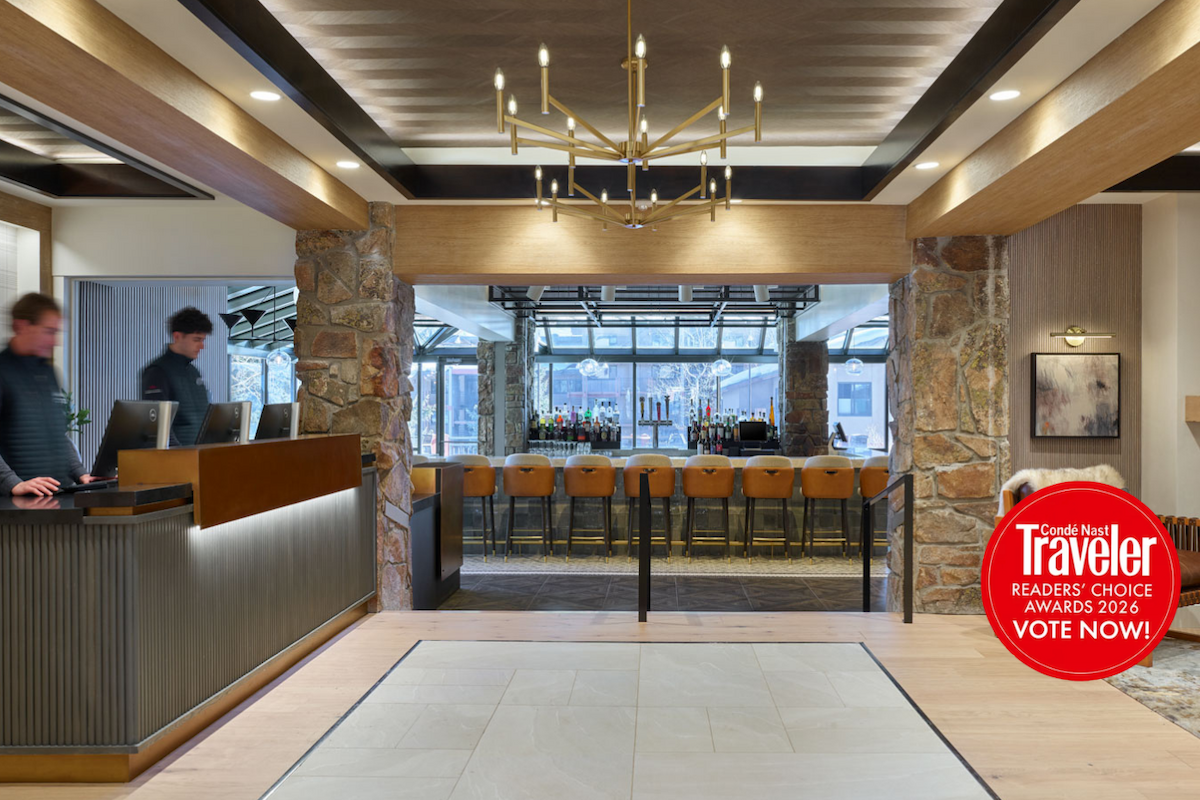 A stylish hotel lobby with a reception desk, stone pillars, warm lighting, and a glassed-in bar area beyond the seating lounge. Vote now.
