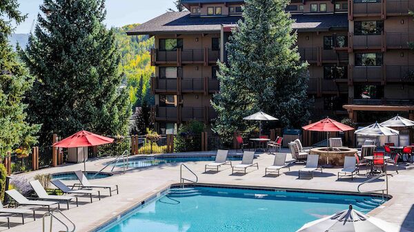 A resort-style pool area with lounge chairs, red umbrellas, and a multi-story brown hotel building surrounded by tall trees, sunny day.