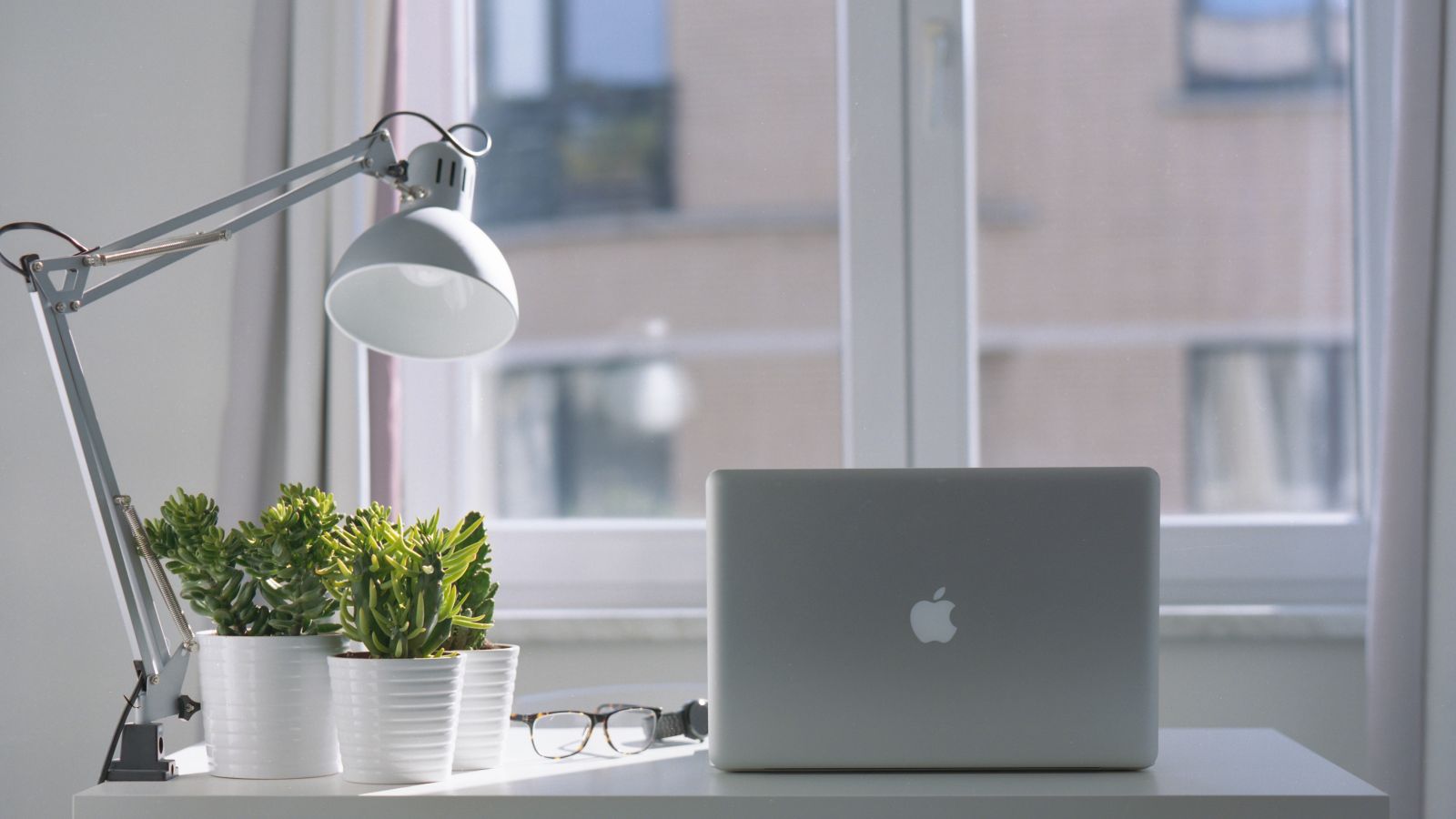 A minimalist desk with a laptop, potted plants, a desk lamp, and eyeglasses is set against a window with a blurred building view outside the window.