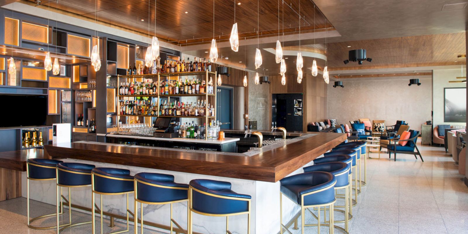 A modern bar with a wooden ceiling, an illuminated back shelf of liquor, pendant lights, and blue cushioned stools surrounding the counter.