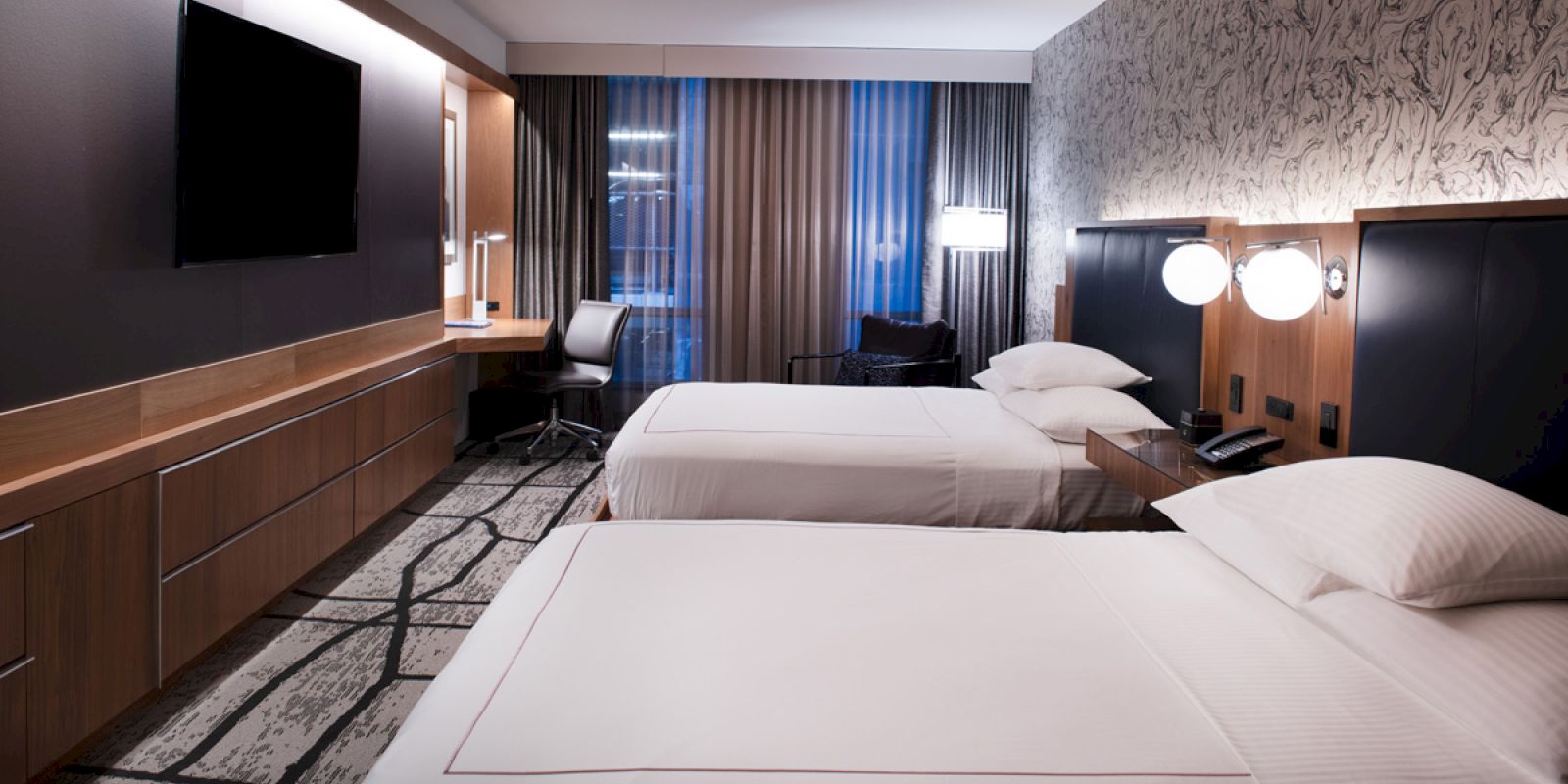The image shows a modern hotel room with two neatly made beds, a wall-mounted TV, a desk with a chair, and contemporary decor with patterned wallpaper.
