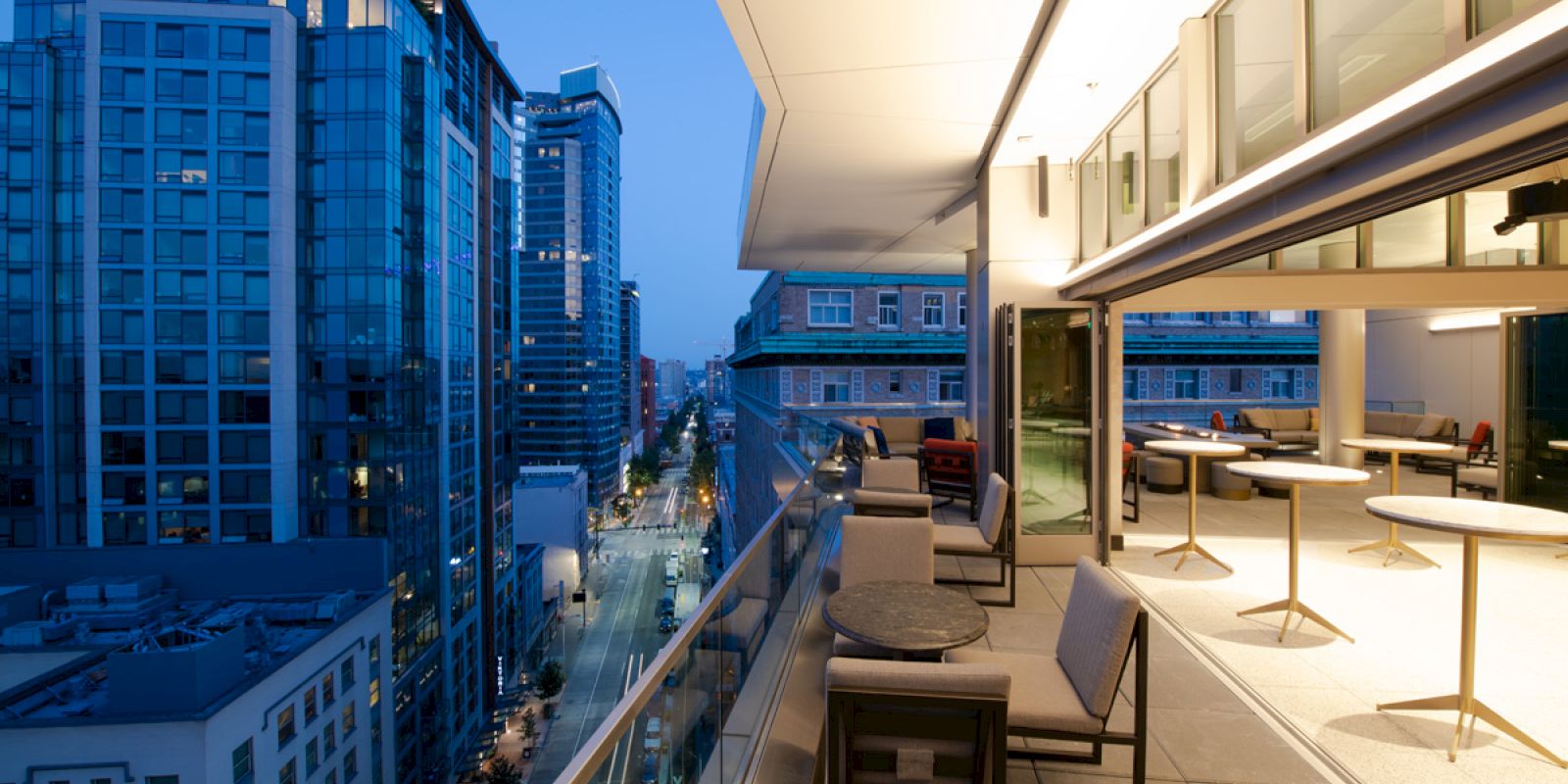 An evening view of a modern urban balcony with seating and tables, overlooking a bustling city street lined with tall buildings.