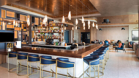 A modern bar with blue bar stools, a well-stocked liquor shelf, overhead lighting, and a seating area with couches in the background.
