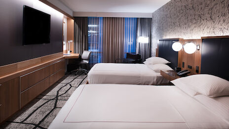 The image shows a modern hotel room with two neatly made beds, a flat-screen TV, a work desk and chair, lamps, and large windows with curtains.