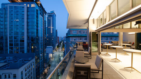 A modern cityscape at dusk with a view from a high balcony featuring outdoor seating and tables. Tall buildings line the street below.