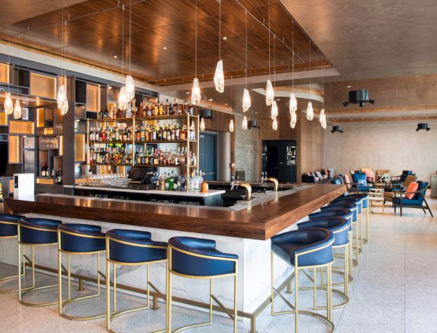 This image shows a modern bar with a sleek wooden counter, blue and gold stools, hanging lights, and a well-stocked liquor shelf in the background.