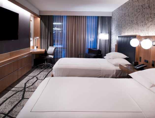 A modern hotel room with two beds, a large TV, a desk and chair by the window, and stylish patterned wallpaper. The room has a clean, contemporary design.