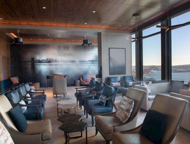 The image shows a modern, stylish lounge with comfortable seating, panoramic windows offering a scenic view, and a cityscape mural on one wall.