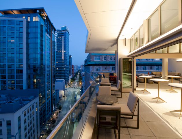 A modern cityscape view from a high-rise building's balcony at dusk, featuring outdoor seating and tables, with city lights beginning to turn on.