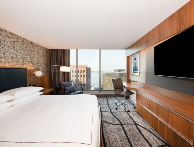 A modern hotel room with a large bed, a wall-mounted TV, a desk with a chair, and a view of city buildings through the window in the background.
