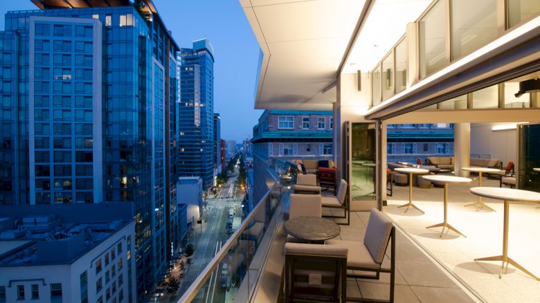 This image shows a modern balcony with seating, overlooking a city street and high-rise buildings, during dusk with the city lights starting to illuminate.