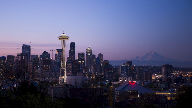 The image shows a city skyline at dusk, featuring the Space Needle, high-rise buildings, and a distant mountain range under a twilight sky.