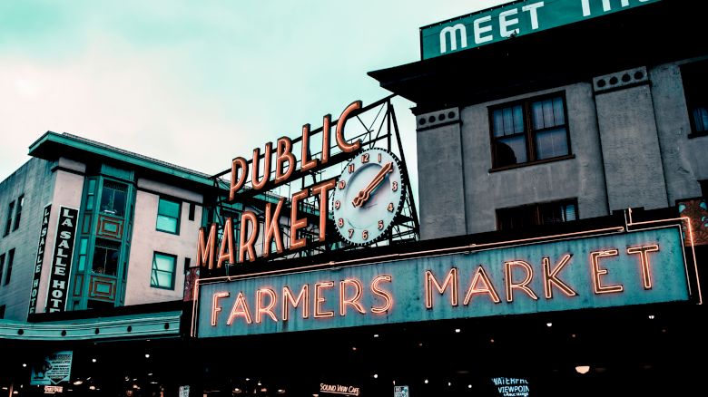 The image shows a neon sign reading "Public Market" and "Farmers Market" with a clock and surrounding buildings at a marketplace.