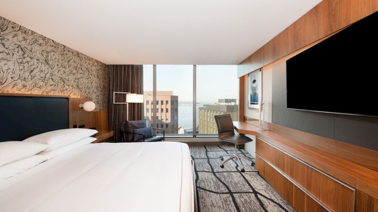 A modern hotel room features a large bed, a desk with a chair, a flat-screen TV, a comfortable chair, and a view of city buildings through large windows.